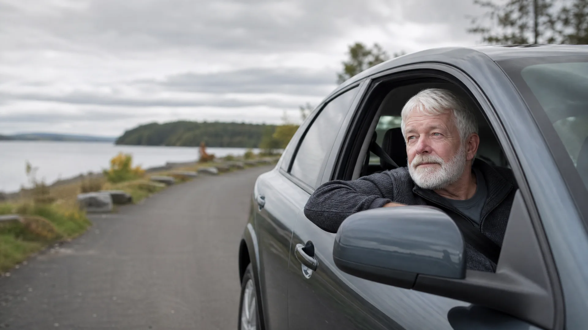 An older man with white hair and a beard rests his arm on the open window of a dark grey car parked on a paved path beside a body of water under an overcast sky.