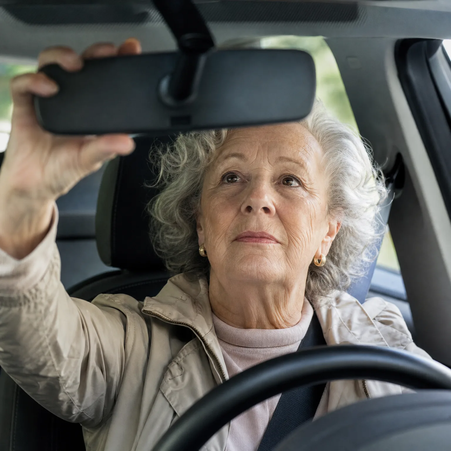An older woman with gray curly hair, wearing a beige jacket and seat belt, reaches up to adjust the rearview mirror while seated behind the steering wheel of a car.
